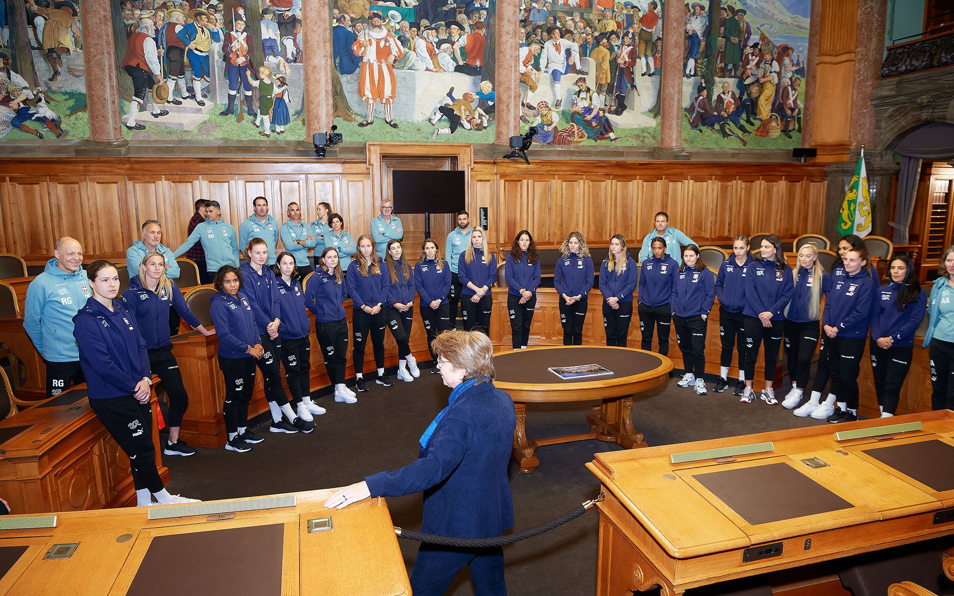 La ministre des sports explique aux joueuses la fresque murale de la salle du Conseil national et leur présente le déroulement des débats dans la salle du Conseil des États.
